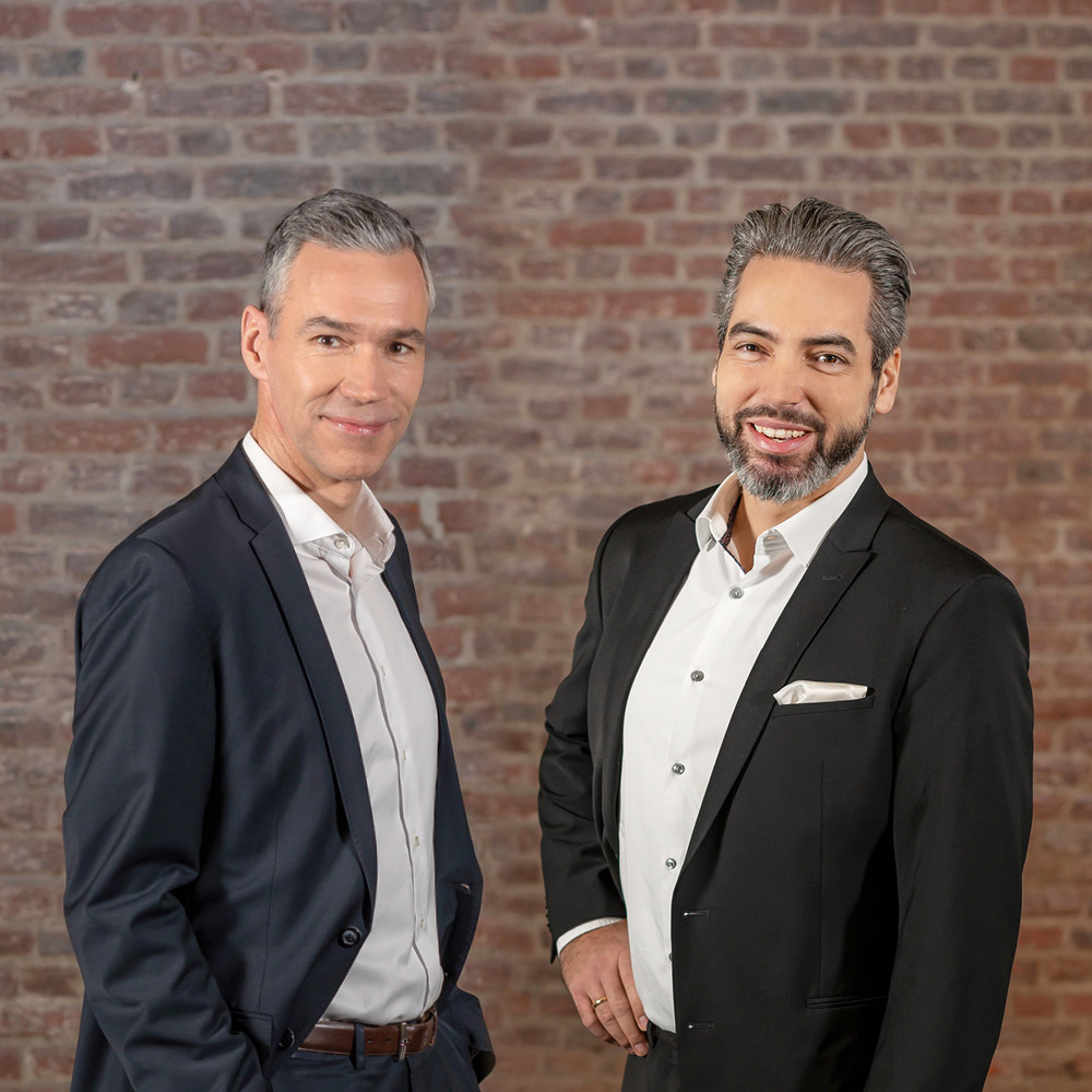 Das Bild zeigt TBF Senior Fondsmanager Patrick Vogel und den TBF Vertriebsdirektor Sven Wiede nebeneinander stehend vor einer Ziegelwand in Bad Homburg